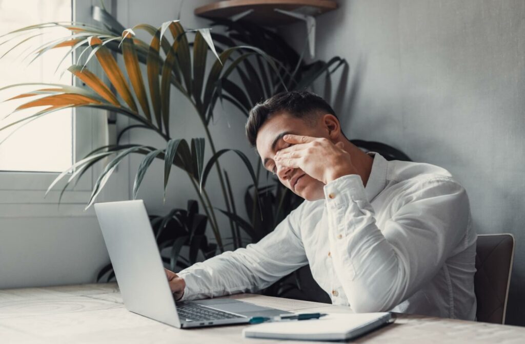 A young adult rubbing their eyes in discomfort and frustration from digital eye strain while trying to work on their laptop.
