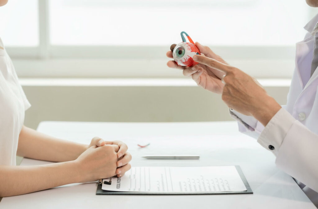 An eye care professional holding and pointing to an anatomical eye model while explaining to a patient during a consultation. A medical form and pen are on the table between them.