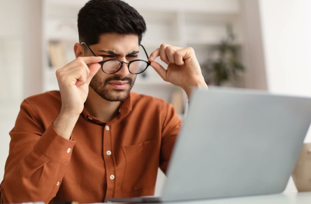 Man squinting and adjusting his glasses while looking at a laptop screen, appearing to experience blurry vision or eye strain.