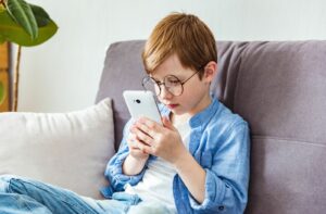 A child wearing glasses sits on a couch holding a smartphone close to their face.