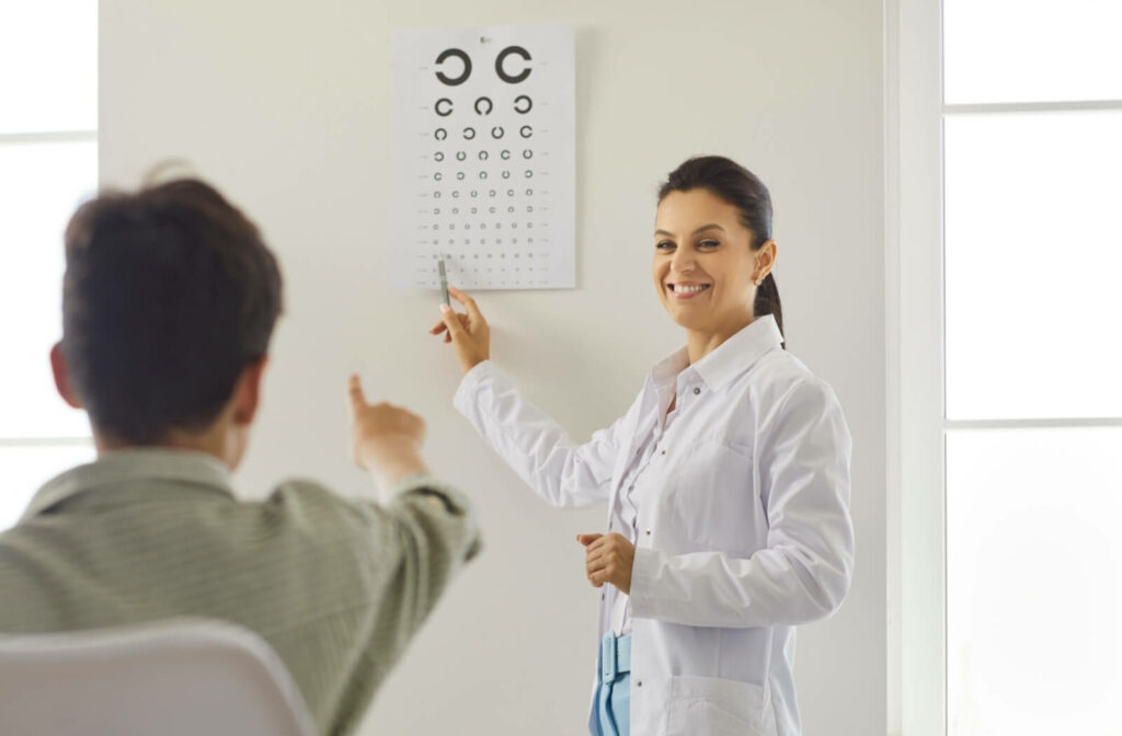 An optometrist points to a vision chart on the wall while smiling at a seated patient who is responding during an eye exam.