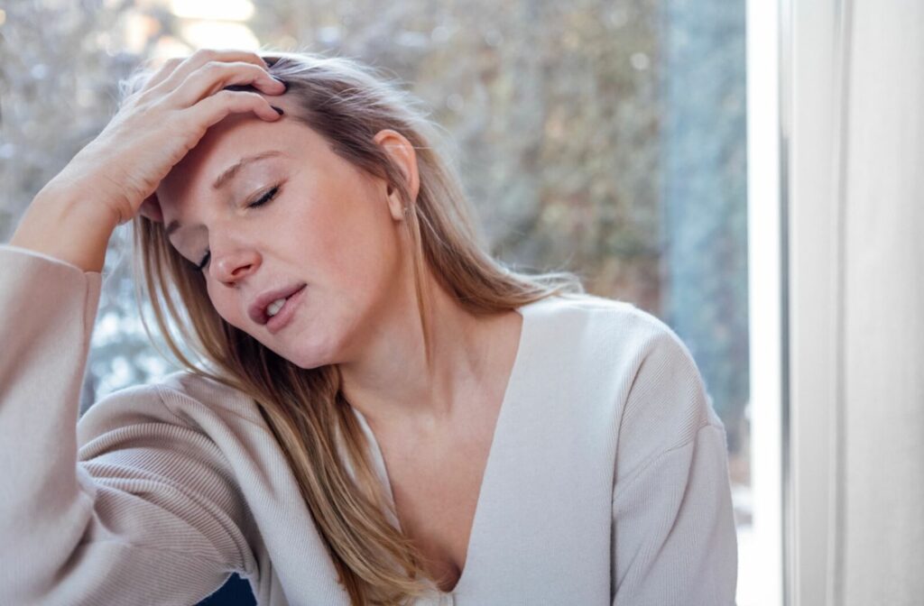 A person sitting indoors with her hand on her forehead, appearing tired or experiencing eye strain, wearing a light sweater with a soft-focus background near a window.