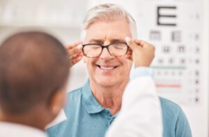 A person smiling as an optometrist helps him try on a pair of eyeglasses during an eye exam, with an eye chart visible in the background.