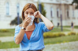 An individual standing outdoors holding a tissue to their nose and rubbing one eye due to allergy symptoms.