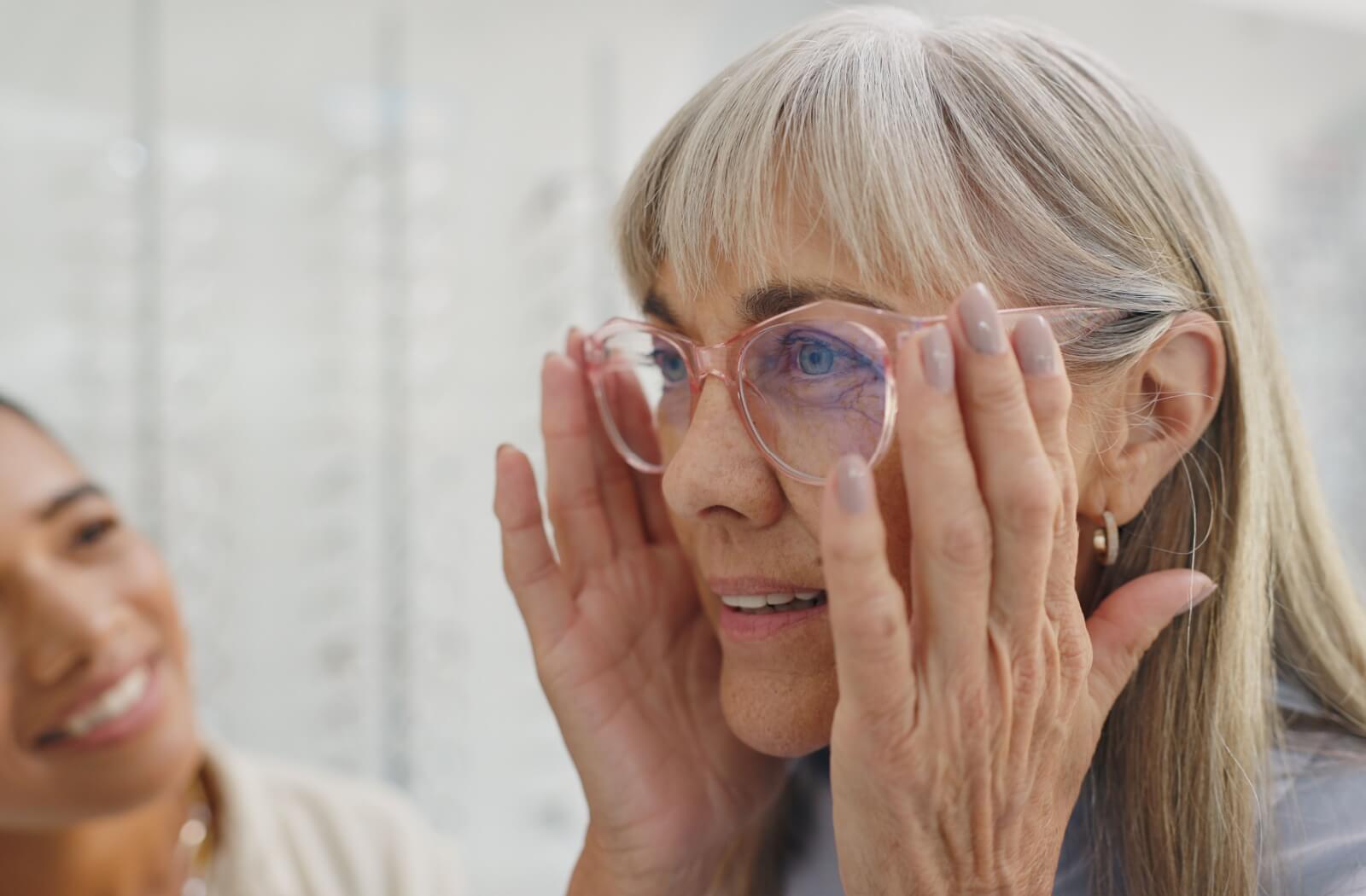 An individual trying on pink-framed eyeglasses while an eye care professional smiles in the background.
