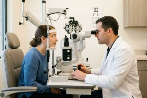 An optometrist examines a patient's eyes using a slit lamp in an exam room, with an eye chart on the wall behind.